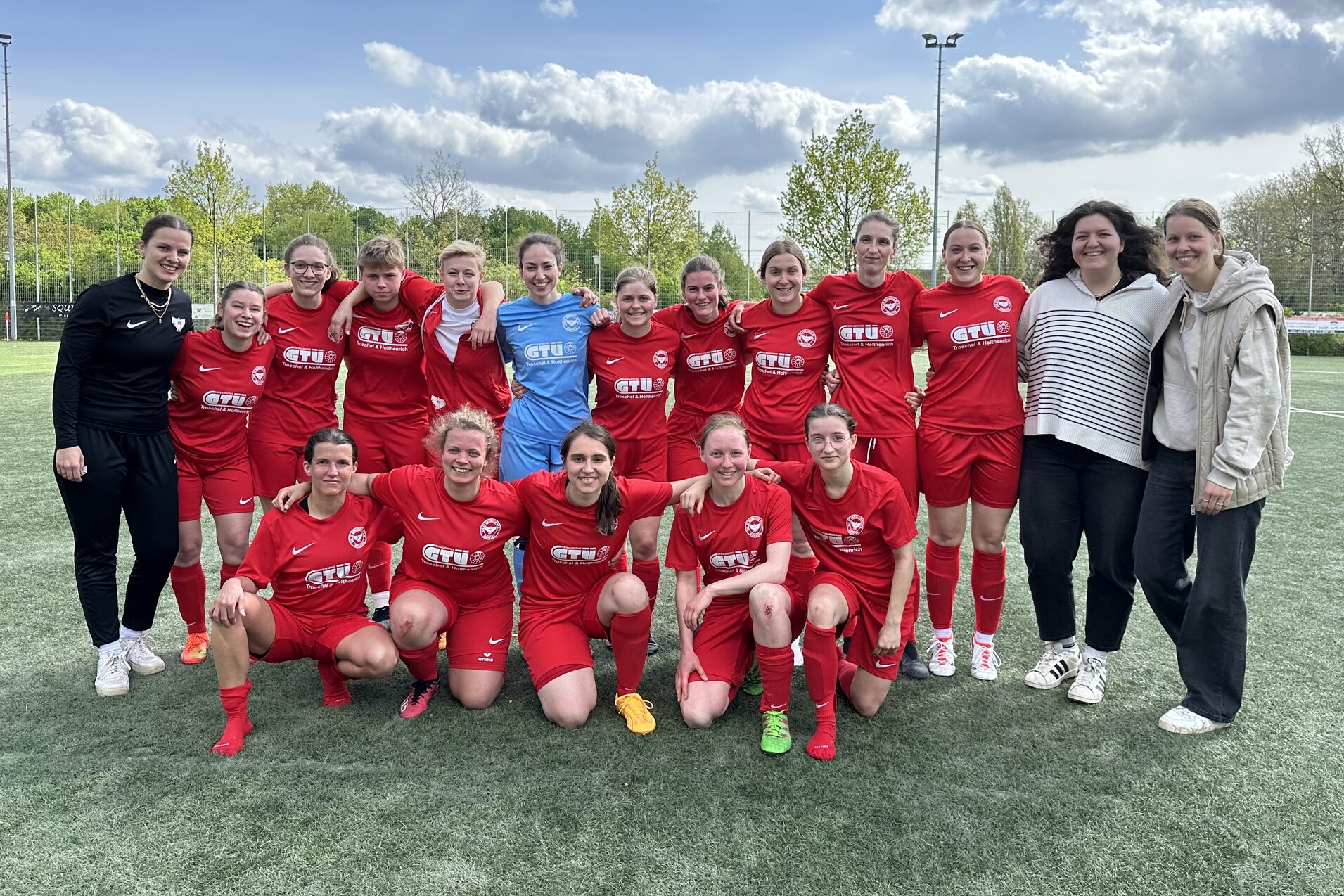 Gruppe von 15 Fu&szlig;ballspielerinnen in roten Trikots und 3 Trainerinnen auf einem Sportplatz, umgeben von B&auml;umen.
