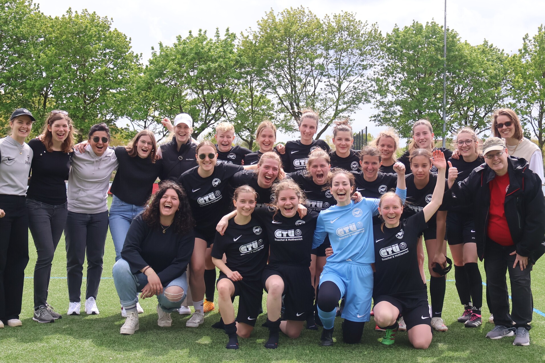 Eine Gruppe von 24 Frauen in schwarzen Fu&szlig;balltrikots posiert l&auml;chelnd auf einem Rasenplatz im Freien.
