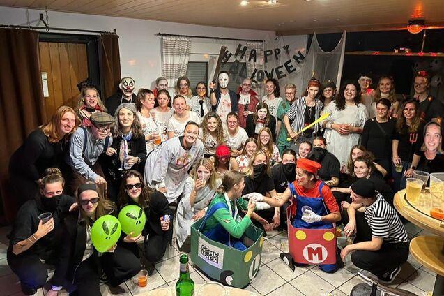 Gruppe von Menschen in Halloween-Kost&uuml;men posiert fr&ouml;hlich vor einer dekorierten Wand mit &bdquo;Happy Halloween&ldquo;-Banner.