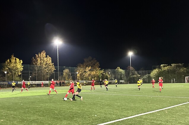 Fu&szlig;ballspiel bei Nacht mit Spielern in roten und gelben Trikots auf einem Kunstrasenplatz unter Flutlicht.