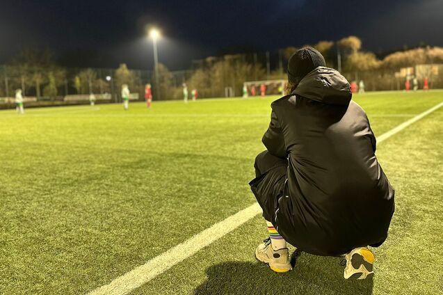 Person im schwarzen Mantel hockt am Rand eines Fu&szlig;ballfeldes, w&auml;hrend im Hintergrund ein Spiel stattfindet.