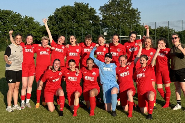 Gruppe von 18 Frauen in roten Fu&szlig;balltrikots posiert fr&ouml;hlich auf dem Rasen, einige heben die Arme zum Sieg.