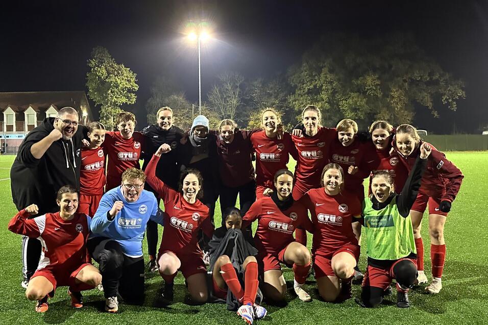 Gruppe von Frauenfu&szlig;ballspielerinnen in roten Trikots posiert nach dem Spiel auf einem beleuchteten Kunstrasenplatz.