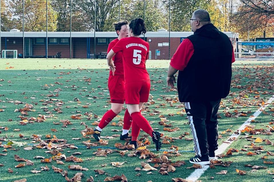 Zwei Fu&szlig;ballspielerinnen in roten Trikots stehen auf einem unebenen Platz mit herbstlichen Bl&auml;ttern, ein Trainer beobachtet sie.