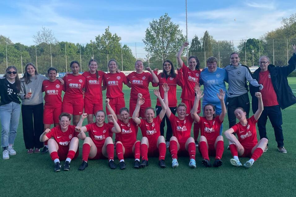 Gruppenfoto eines Frauenfu&szlig;ballteams in roten Trikots, l&auml;chelnd und mit erhobenen H&auml;nden auf einem Sportplatz.