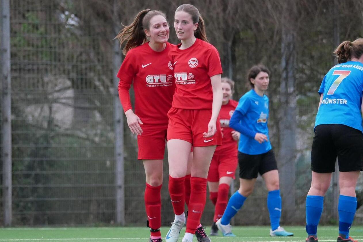 Zwei Fu&szlig;ballspielerinnen in roten Trikots feiern einen Erfolg auf dem Spielfeld, w&auml;hrend andere Spielerinnen im Hintergrund stehen.