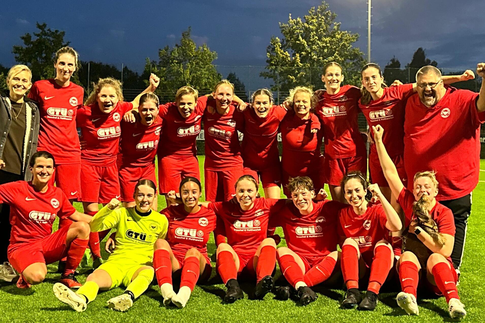 Gruppe von 16 Fu&szlig;ballspielerinnen in roten Trikots posiert fr&ouml;hlich nach einem Spiel auf einem beleuchteten Rasenplatz.