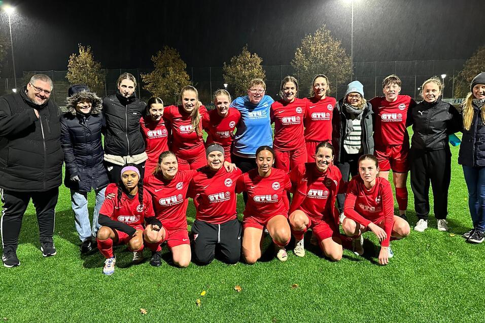 Gruppe von 18 Frauen und M&auml;nnern in Sportkleidung auf einem Fu&szlig;ballplatz bei Nacht, umgeben von Flutlichtern.