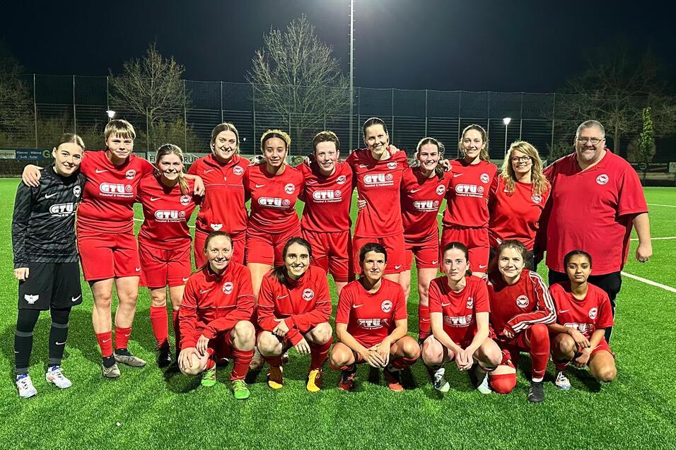 Eine Gruppe von 15 Fu&szlig;ballspielerinnen in roten Trikots posiert zusammen mit zwei Trainern auf einem beleuchteten Spielfeld.