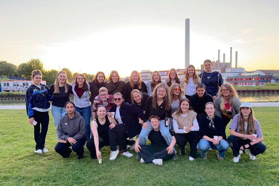 Gruppe von jungen Frauen und M&auml;nnern posiert l&auml;chelnd auf einer Wiese vor einem Sonnenuntergang und einem Industriegeb&auml;ude.