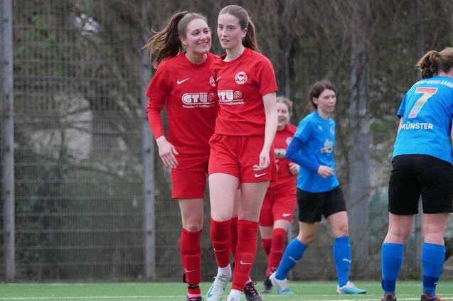 Zwei Fu&szlig;ballspielerinnen in roten Trikots feiern einen Erfolg auf dem Spielfeld, w&auml;hrend andere Spielerinnen im Hintergrund stehen.