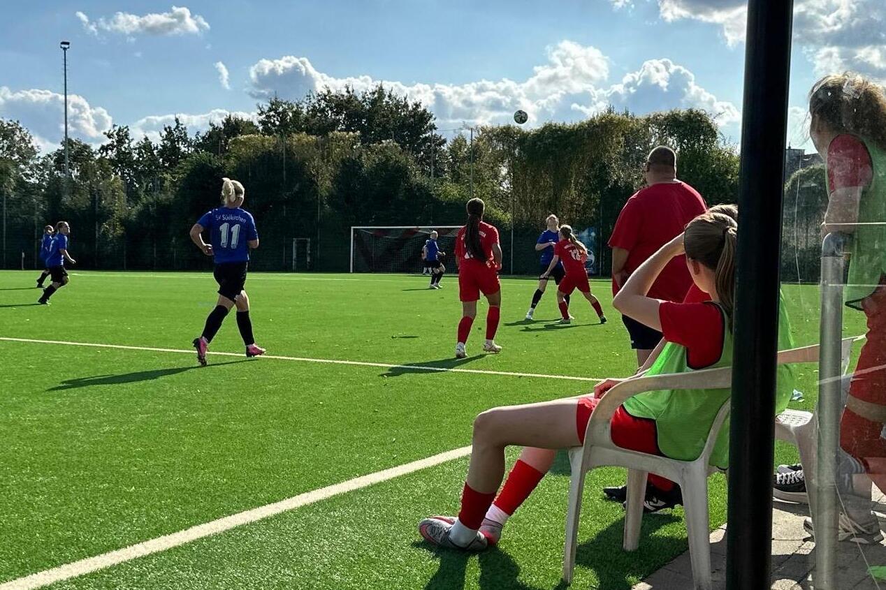 Fu&szlig;ballspiel auf Kunstrasen mit Spielerinnen in roten und blauen Trikots und Zuschauern auf der Bank.