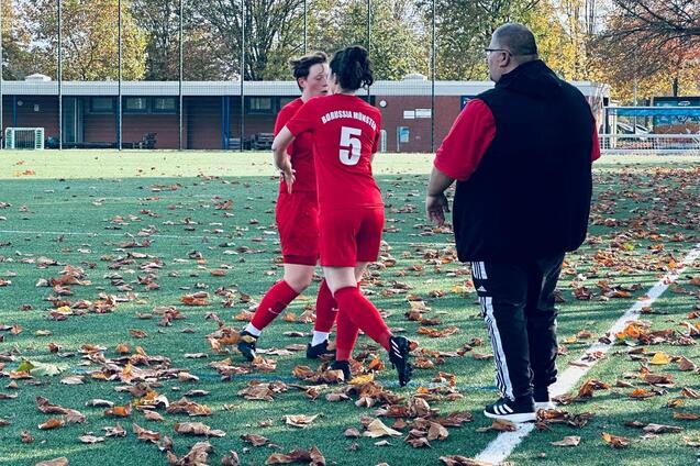 Zwei Fu&szlig;ballspielerinnen in roten Trikots stehen auf einem unebenen Platz mit herbstlichen Bl&auml;ttern, ein Trainer beobachtet sie.