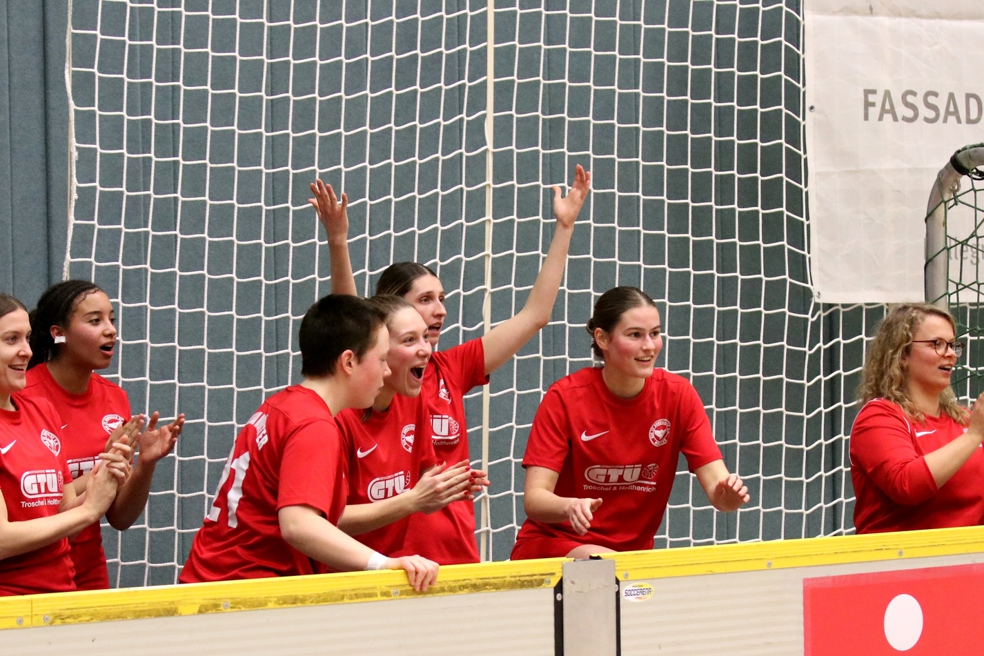 Gruppe jubelnder Frauen in roten Trikots am Spielfeldrand mit Netz im Hintergrund.