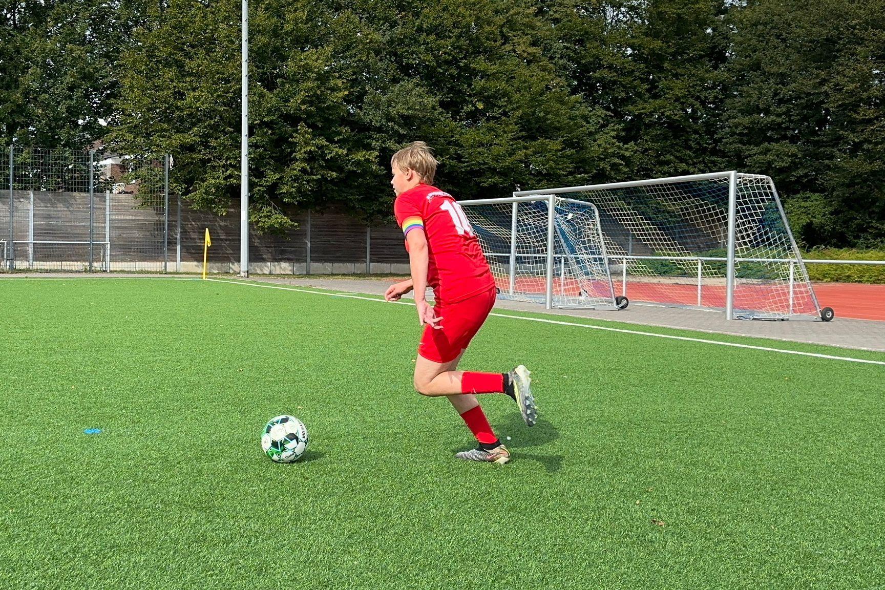 Fu&szlig;ballspieler in rotem Trikot sprintet &uuml;ber ein Kunstrasenfeld in Richtung Ball. Im Hintergrund stehen Tore.