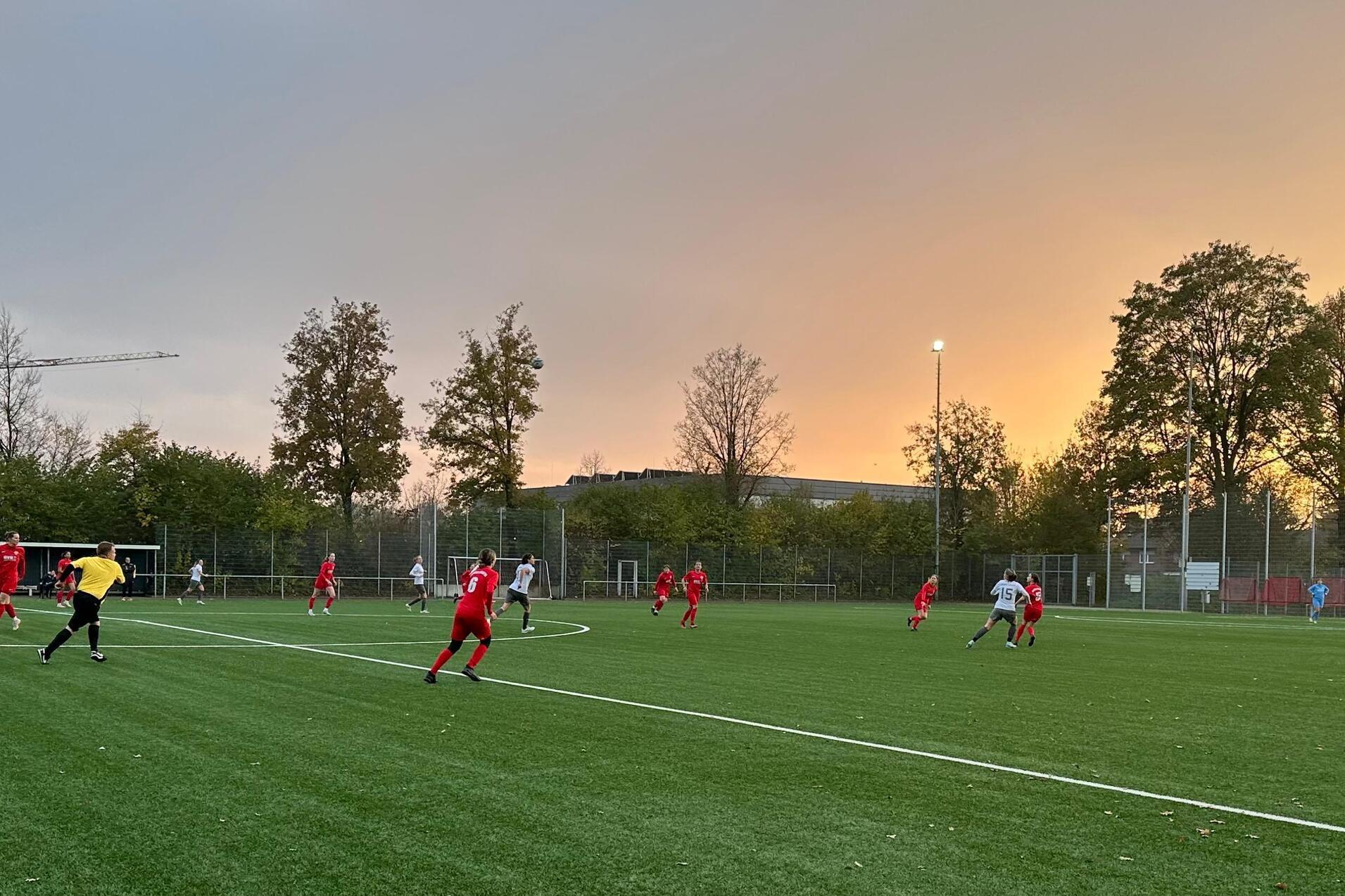 Fu&szlig;ballspiel auf einem Kunstrasenplatz bei Sonnenuntergang, Spieler in roten und wei&szlig;en Trikots in Aktion.