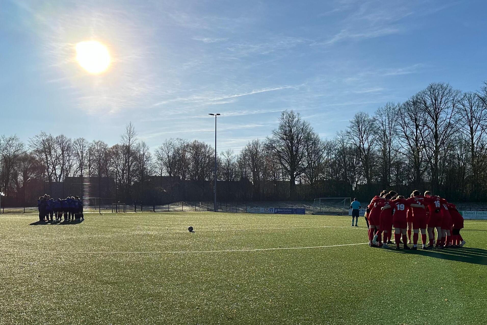 Zwei Fu&szlig;ballmannschaften in ihren Trikots stehen sich auf einem Rasenplatz gegen&uuml;ber, Sonnenlicht am Himmel.