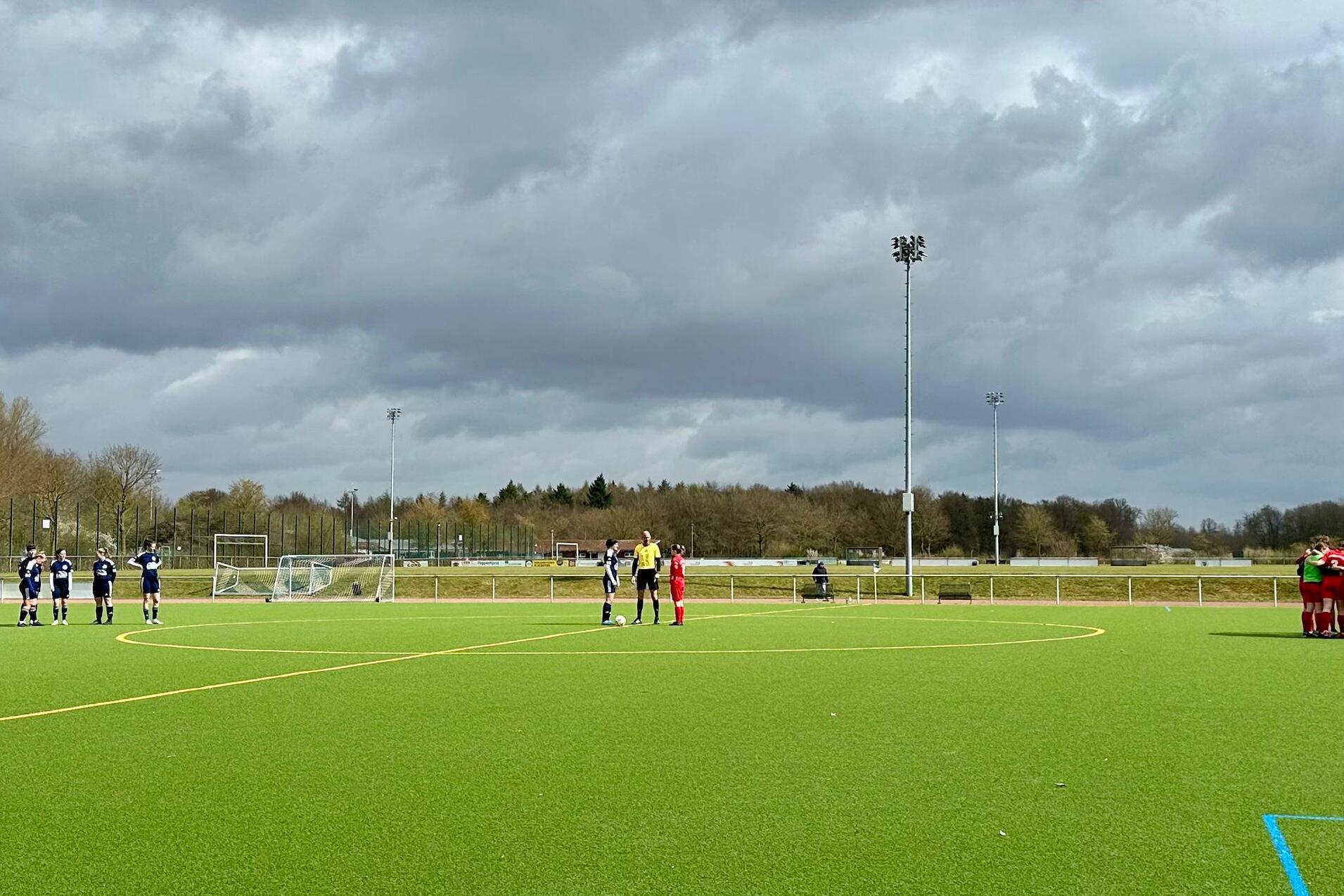 Zwei Fu&szlig;ballmannschaften stehen sich auf einem Kunstrasenplatz gegen&uuml;ber, unter einem bew&ouml;lkten Himmel.