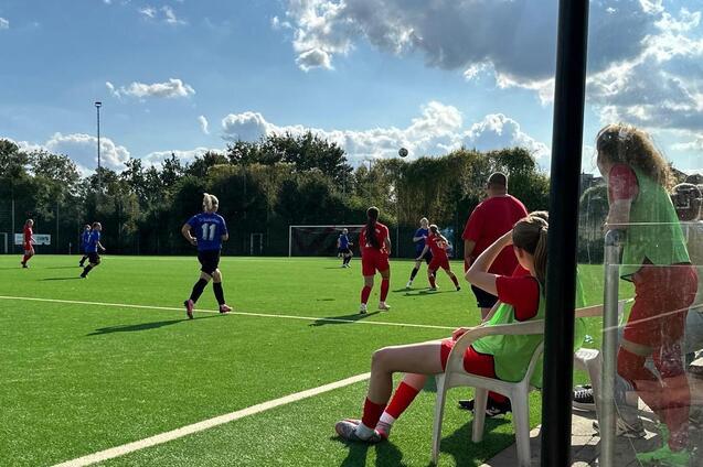 Fu&szlig;ballspiel auf Kunstrasen mit Spielerinnen in roten und blauen Trikots und Zuschauern auf der Bank.