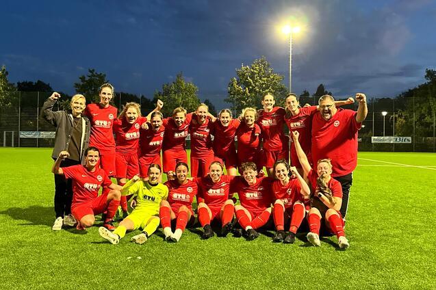Gruppe von 16 Fu&szlig;ballspielerinnen in roten Trikots posiert fr&ouml;hlich nach einem Spiel auf einem beleuchteten Rasenplatz.