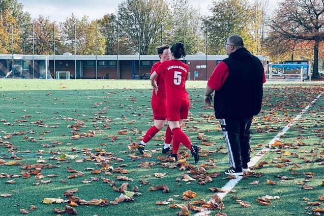 Zwei Fu&szlig;ballspielerinnen in roten Trikots stehen auf einem unebenen Platz mit herbstlichen Bl&auml;ttern, ein Trainer beobachtet sie.