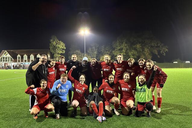 Gruppe von Frauenfu&szlig;ballspielerinnen in roten Trikots posiert nach dem Spiel auf einem beleuchteten Kunstrasenplatz.