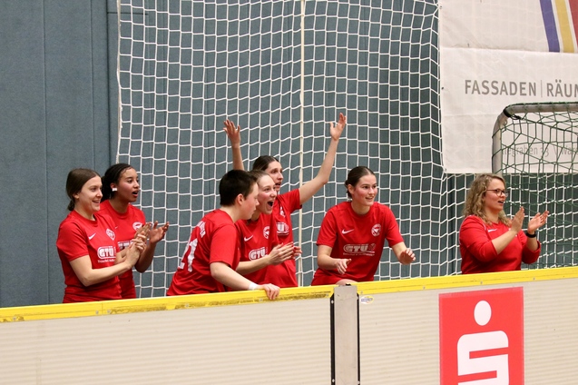Gruppe jubelnder Frauen in roten Trikots am Spielfeldrand mit Netz im Hintergrund.