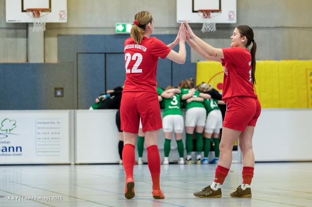 Zwei Fu&szlig;ballspielerinnen in roten Trikots klatschen sich ab, w&auml;hrend im Hintergrund ein Team in gr&uuml;nen Trikots zusammensteht.