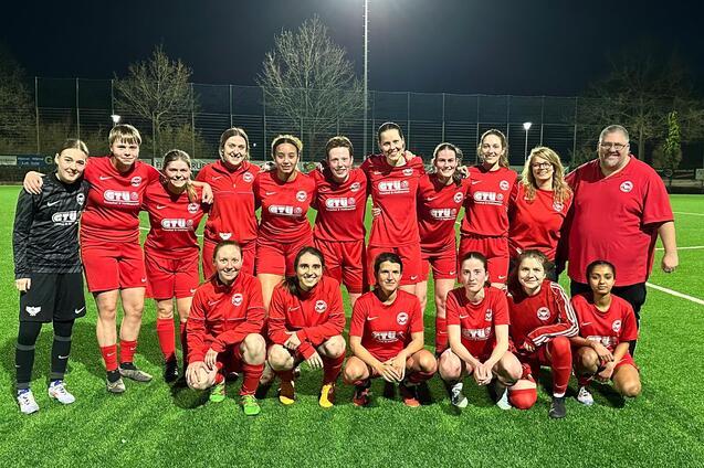 Eine Gruppe von 15 Fu&szlig;ballspielerinnen in roten Trikots posiert zusammen mit zwei Trainern auf einem beleuchteten Spielfeld.