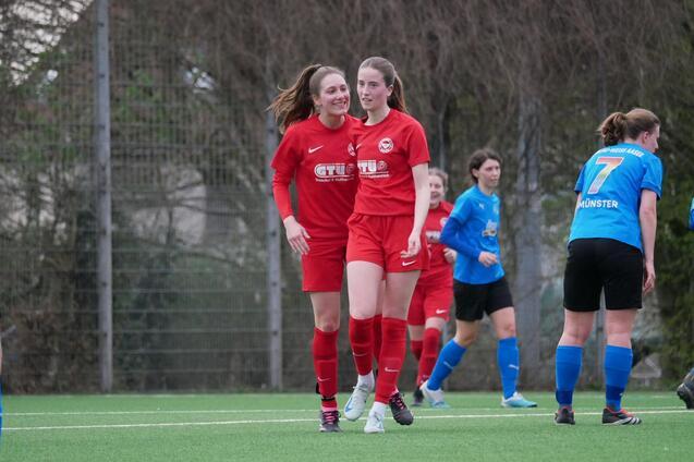 Zwei Fu&szlig;ballspielerinnen in roten Trikots feiern einen Erfolg auf dem Spielfeld, w&auml;hrend andere Spielerinnen im Hintergrund stehen.