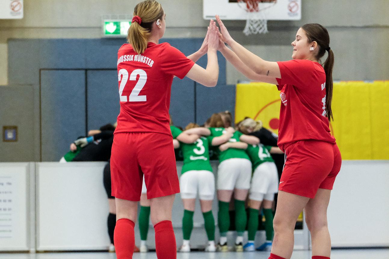 Zwei Fu&szlig;ballspielerinnen in roten Trikots klatschen sich ab, w&auml;hrend im Hintergrund ein Team in gr&uuml;nen Trikots zusammensteht.