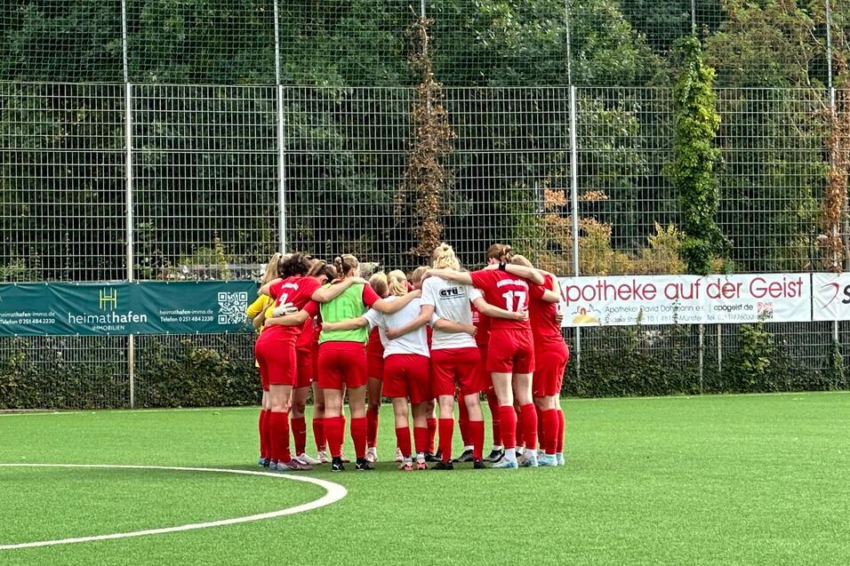Eine Frauenfu&szlig;ballmannschaft in roten Trikots steht im Kreis auf einem Kunstrasenplatz und bespricht sich.