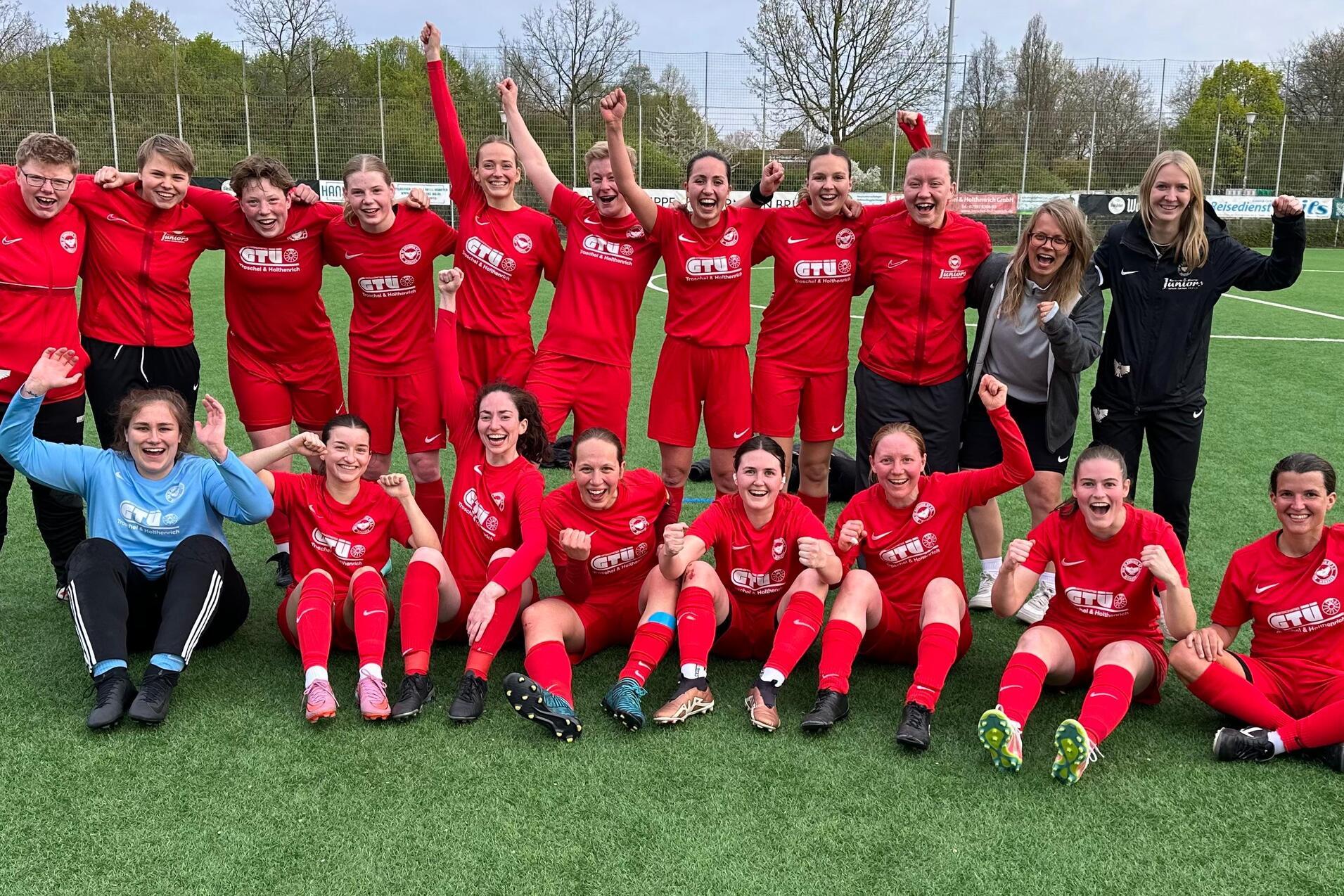 Gruppenselfie von 18 Frauen in roten Fu&szlig;balltrikots, die auf einem Platz jubeln und die Arme in die Luft strecken.
