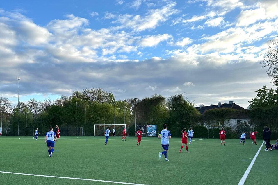 Fu&szlig;ballspiel auf einem Kunstrasenplatz, Spieler in roten und blauen Trikots, B&auml;ume und Wolken im Hintergrund.