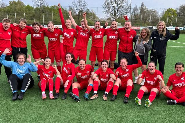 Gruppenselfie von 18 Frauen in roten Fu&szlig;balltrikots, die auf einem Platz jubeln und die Arme in die Luft strecken.