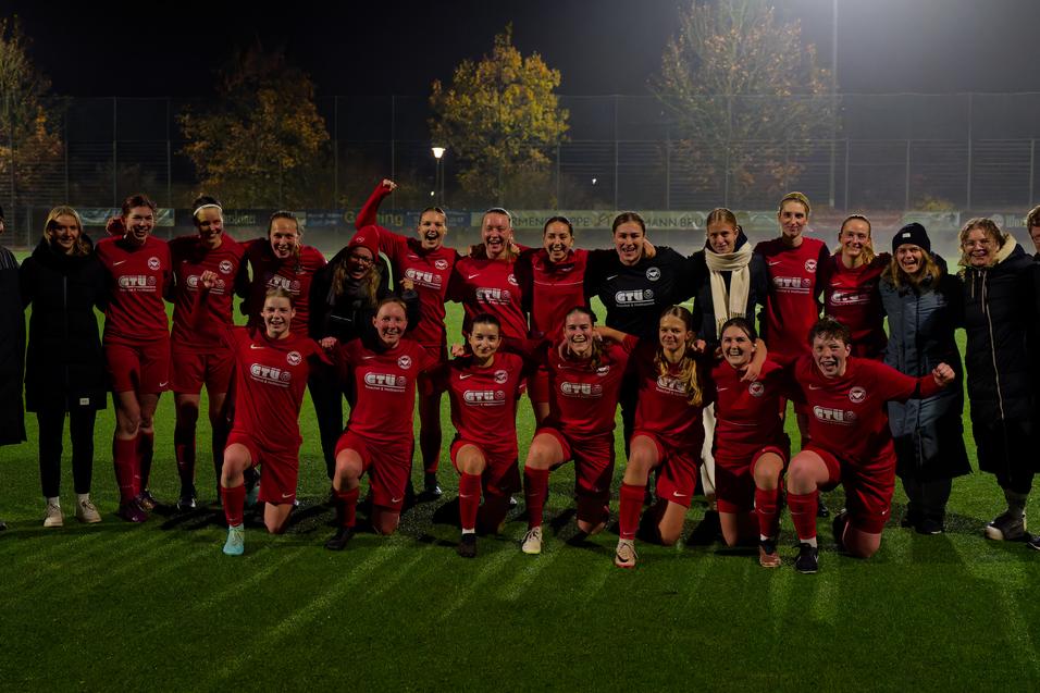 Gruppe von Fu&szlig;ballspielerinnen in roten Trikots posiert jubelnd auf einem gut beleuchteten Kunstrasenplatz.