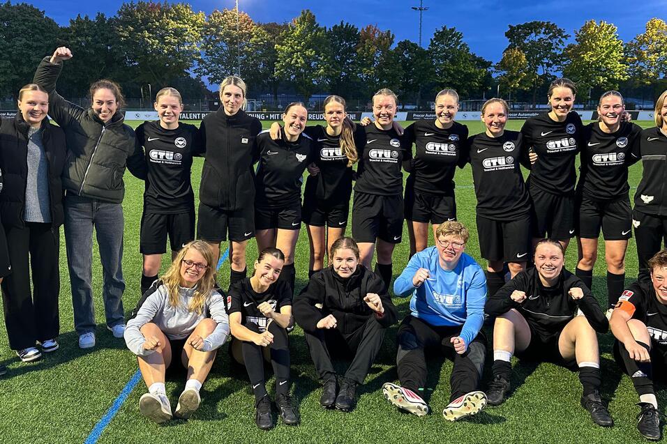 Gruppenshot des Frauenfu&szlig;ballteams in schwarzen Trikots, l&auml;chelnd auf einem Sportplatz unter Abendbeleuchtung.