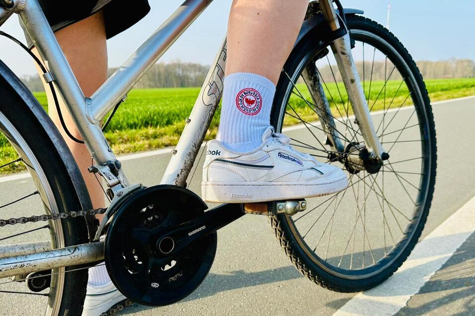 Nahaufnahme eines Fahrradpedals mit einem Fu&szlig;, der in einem wei&szlig;en Schuh und Socke auf einer Radwegfl&auml;che steht.