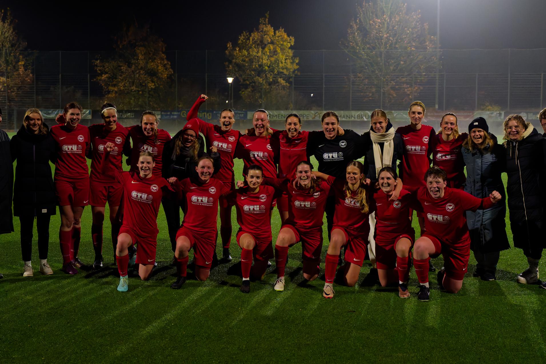 Gruppe von Fu&szlig;ballspielerinnen in roten Trikots posiert jubelnd auf einem gut beleuchteten Kunstrasenplatz.