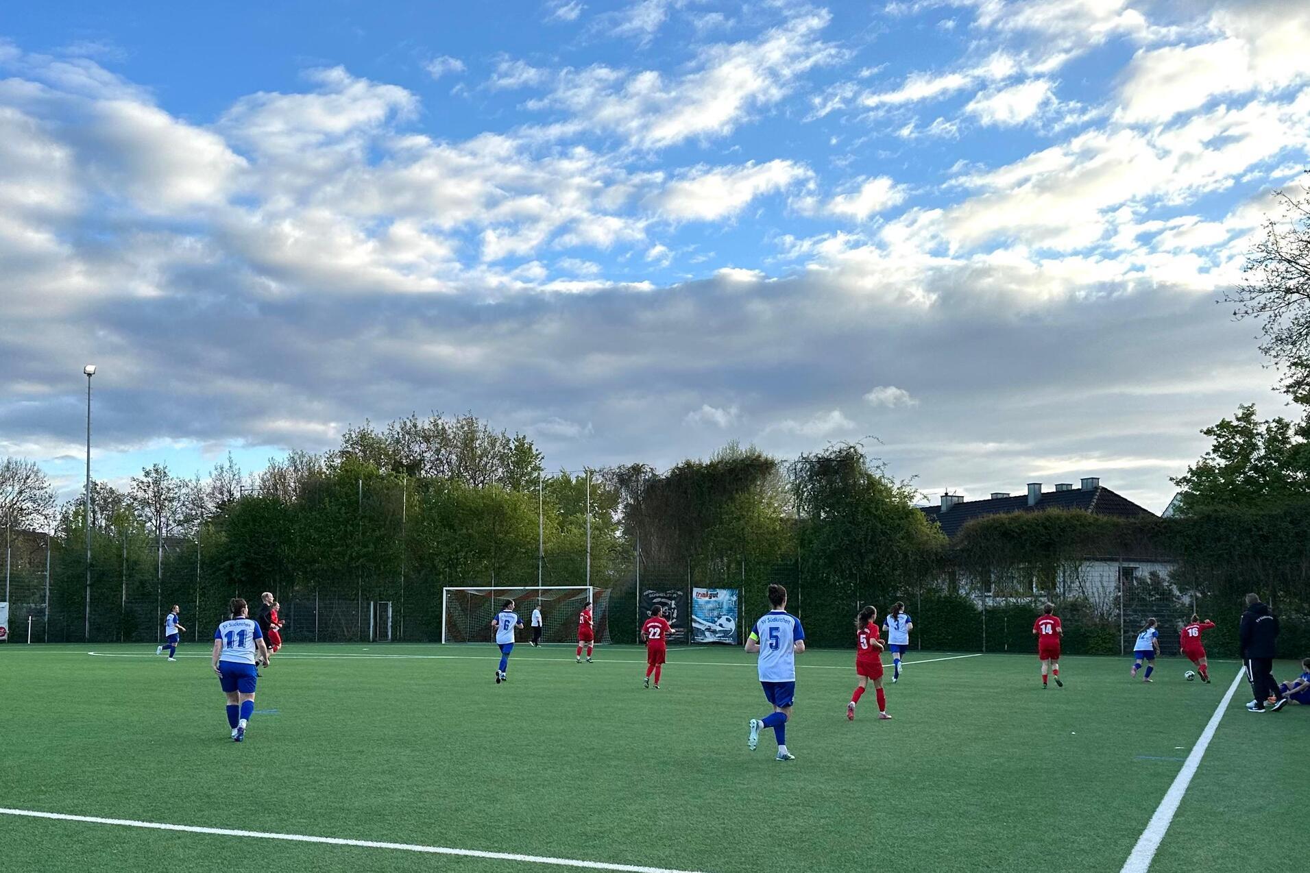 Fu&szlig;ballspiel auf einem Kunstrasenplatz, Spieler in roten und blauen Trikots, B&auml;ume und Wolken im Hintergrund.