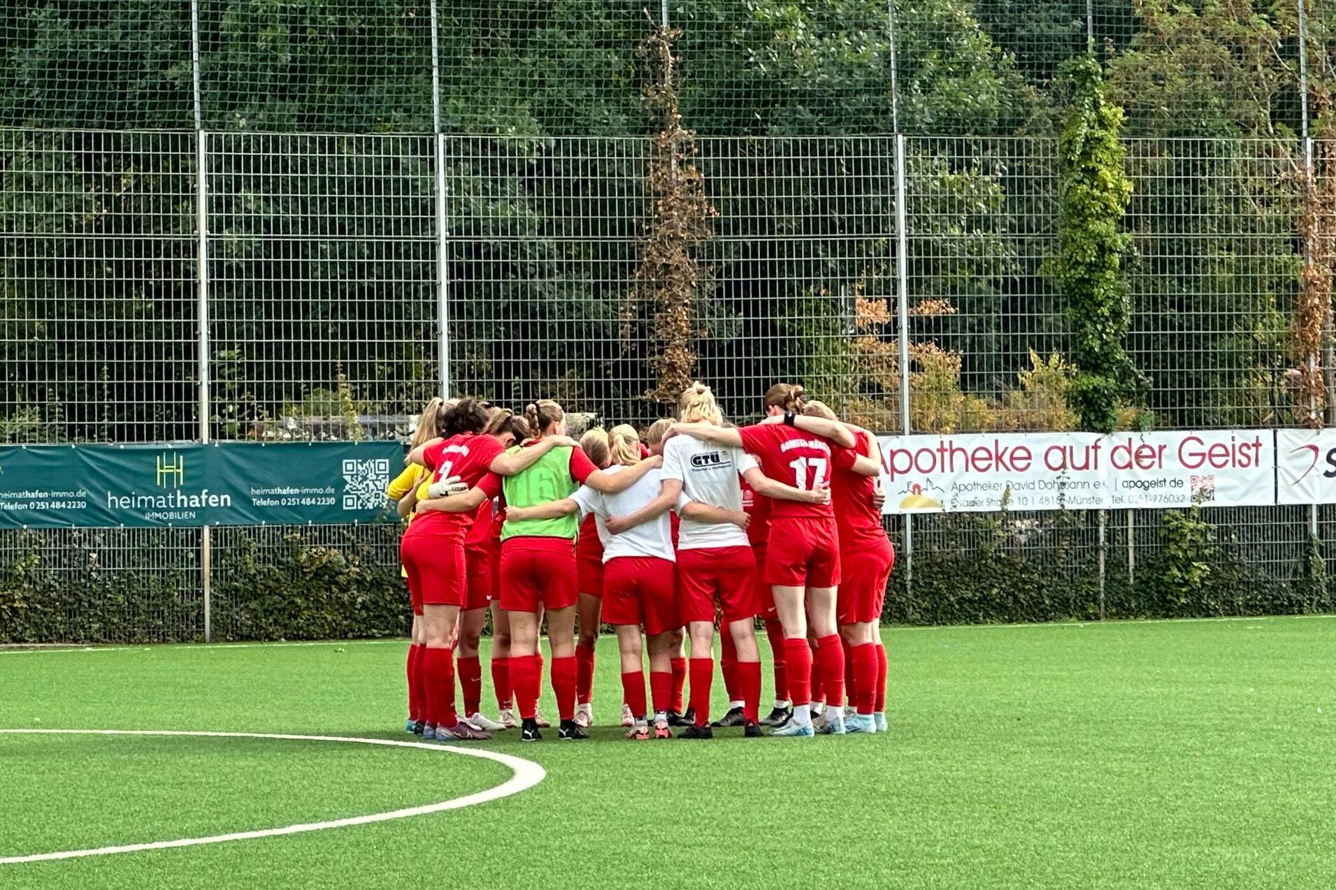 Eine Frauenfu&szlig;ballmannschaft in roten Trikots steht im Kreis auf einem Kunstrasenplatz und bespricht sich.