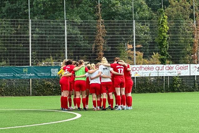 Eine Frauenfu&szlig;ballmannschaft in roten Trikots steht im Kreis auf einem Kunstrasenplatz und bespricht sich.