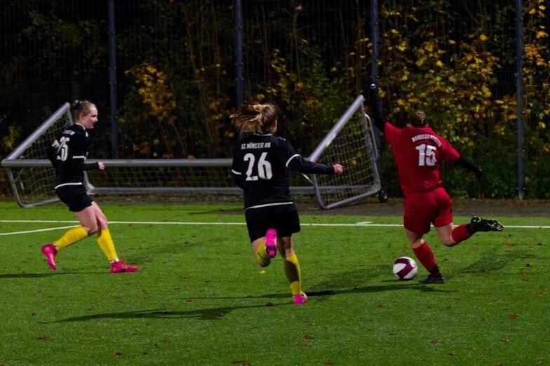Zwei Spielerinnen in schwarzen Trikots verfolgen eine gegnerische Spielerin in rotem Trikot auf einem Fu&szlig;ballplatz.