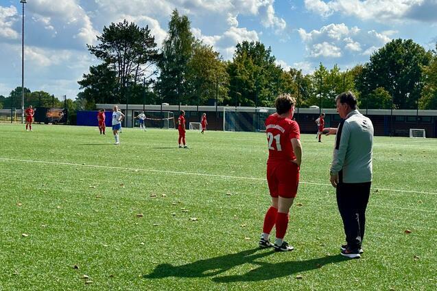 Trainer spricht mit Spielerin in rotem Trikot auf einem Fu&szlig;ballplatz, im Hintergrund sind weitere Spieler aktiv.