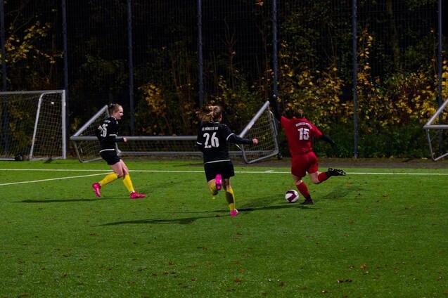 Zwei Spielerinnen in schwarzen Trikots verfolgen eine gegnerische Spielerin in rotem Trikot auf einem Fu&szlig;ballplatz.