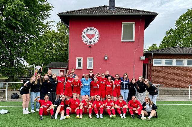 Gruppenfoto einer Fu&szlig;ballmannschaft in roten Trikots vor einem roten Vereinsgeb&auml;ude, umgeben von B&auml;umen.