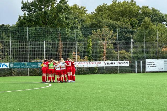 Eine Frauenfu&szlig;ballmannschaft in roten Trikots steht im Kreis auf einem Kunstrasenplatz und bespricht sich.
