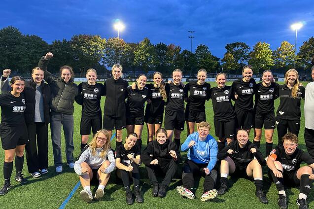 Gruppenshot des Frauenfu&szlig;ballteams in schwarzen Trikots, l&auml;chelnd auf einem Sportplatz unter Abendbeleuchtung.