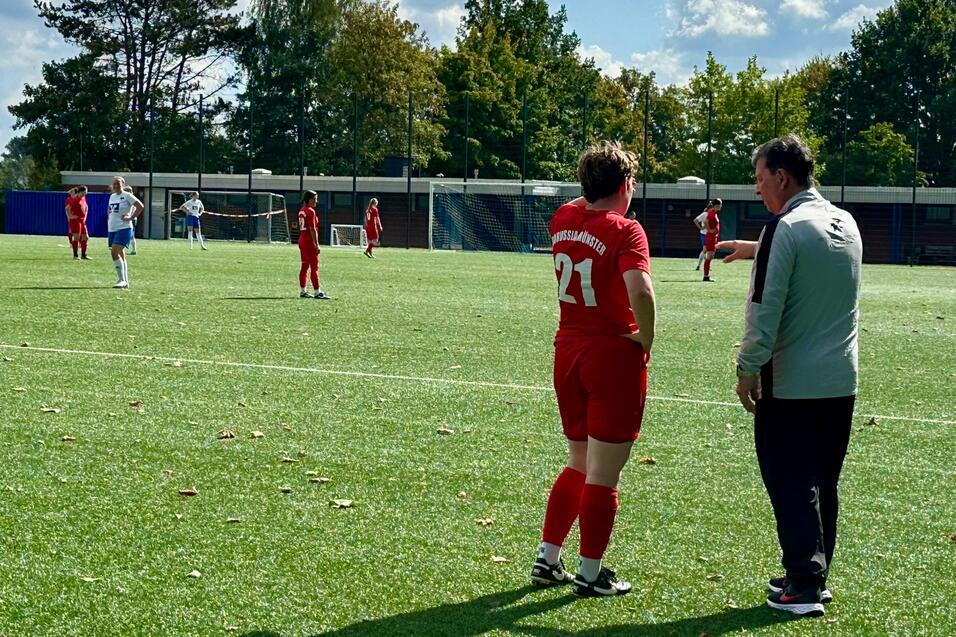 Trainer spricht mit Spielerin in rotem Trikot auf einem Fu&szlig;ballplatz, im Hintergrund sind weitere Spieler aktiv.
