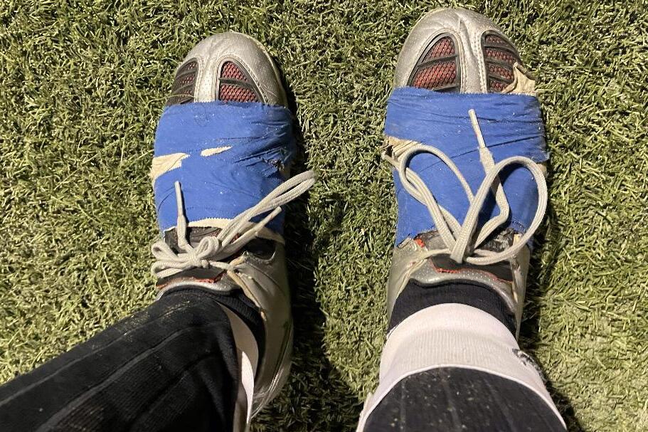 Nahaufnahme von F&uuml;&szlig;en in Fu&szlig;ballschuhen, mit blauer Klebebandreparatur, auf einem Kunstrasenplatz.